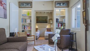 A cozy living room with a sofa, armchair, and a coffee table with a bowl of nuts and a vase of flowers. There are bookshelves with books and decorative items, and a painting on the wall. - Ca' Minore Secret Garden (Venice)
