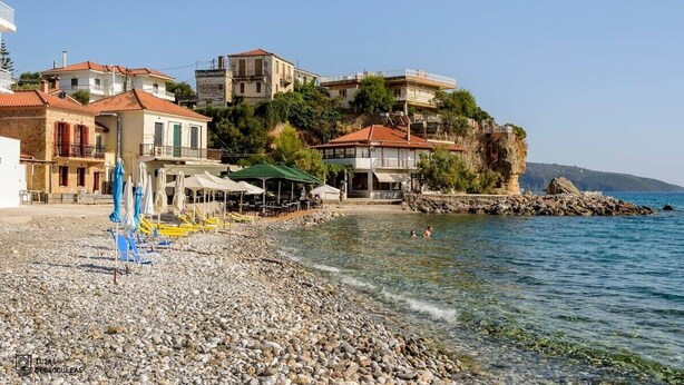 A coastal area with buildings on a hillside overlooking the sea, a pebble beach, and outdoor seating with umbrellas. - Palaiochora Retreat - Sea Sunset Rooftop (Avia Kalamata)