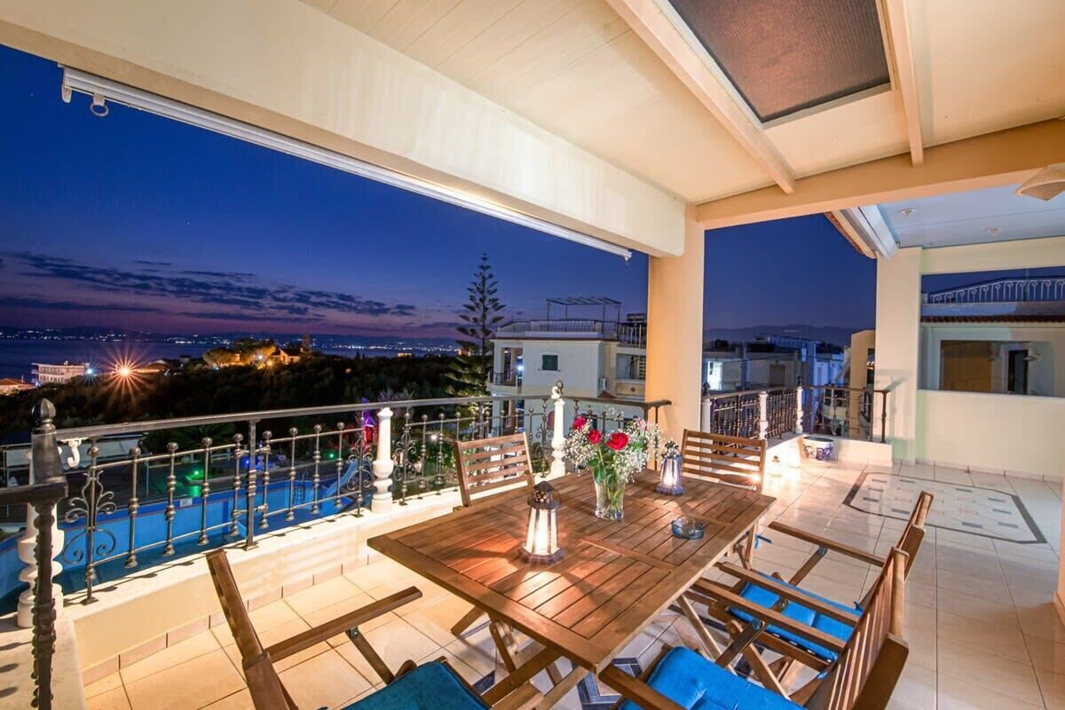 A terrace with a wooden table and chairs, a railing, and a view of the city at dusk.