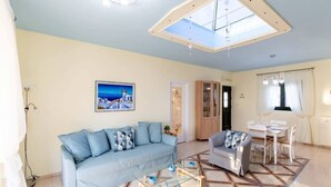 A living room with a sofa, a coffee table, a dining table with chairs, and a ceiling light fixture. - Palaiochora Retreat - Sea Sunset Rooftop (Avia Kalamata)