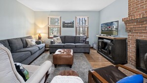 Ein Wohnzimmer mit grauem Sofa, braunem Hocker, Fernseher auf einem Schrank und Kamin.