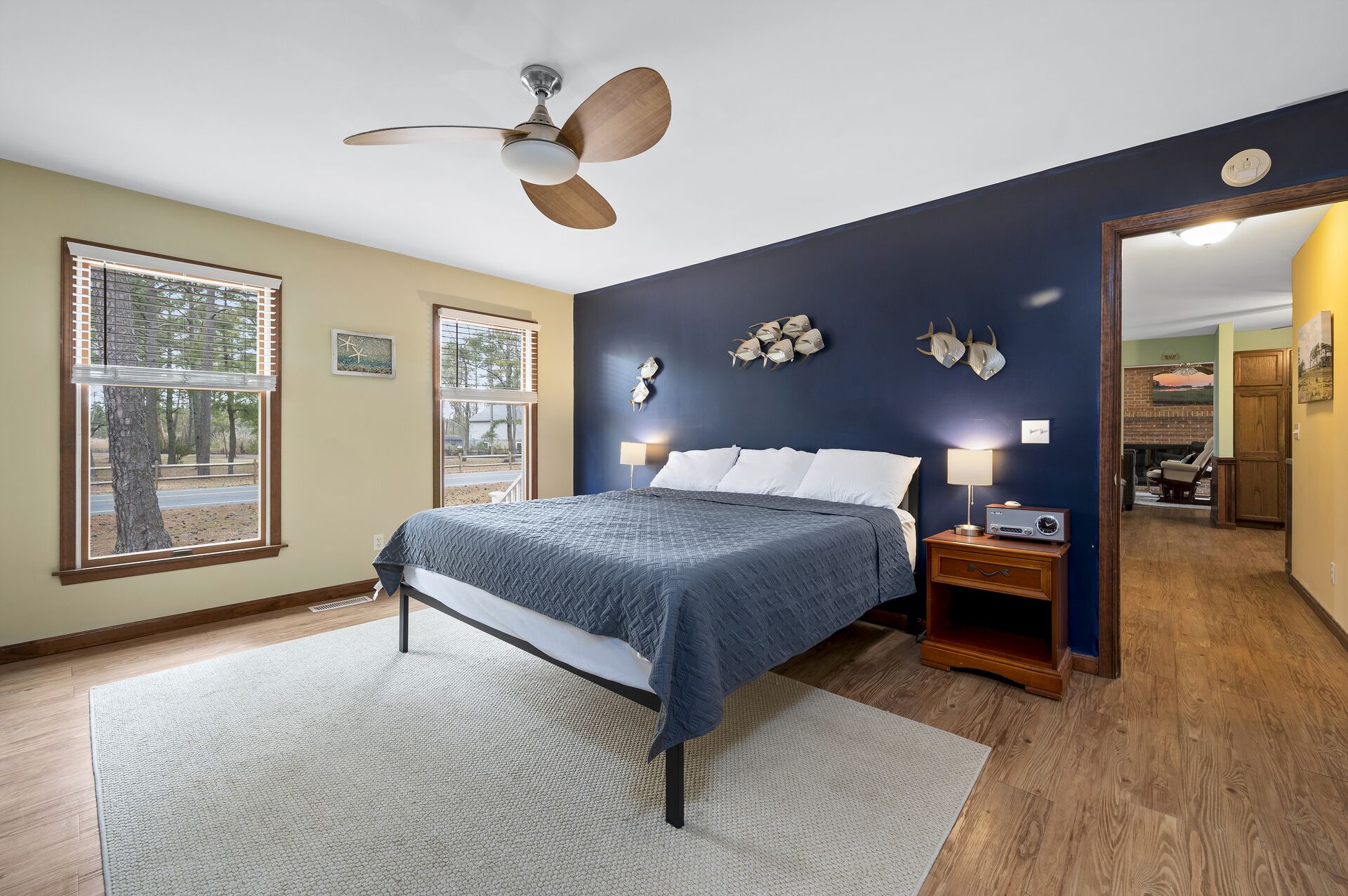 A bedroom with a bed, a ceiling fan, a nightstand with a lamp, and a view of the outdoors through a window.