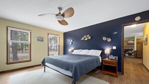 Ein Schlafzimmer mit einem Bett, einem Deckenventilator, einem Nachttisch mit Lampe und Blick ins Freie durch ein Fenster.