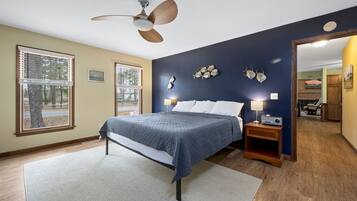 A bedroom with a bed, a ceiling fan, a nightstand with a lamp, and a view of the outdoors through a window.