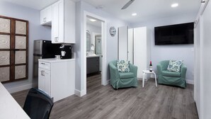 A modern living room with a kitchen area, a dining table, two green armchairs, a small white table, and a flat-screen TV mounted on the wall. - Charming Private Mission Bay Studio 2 (San Diego)