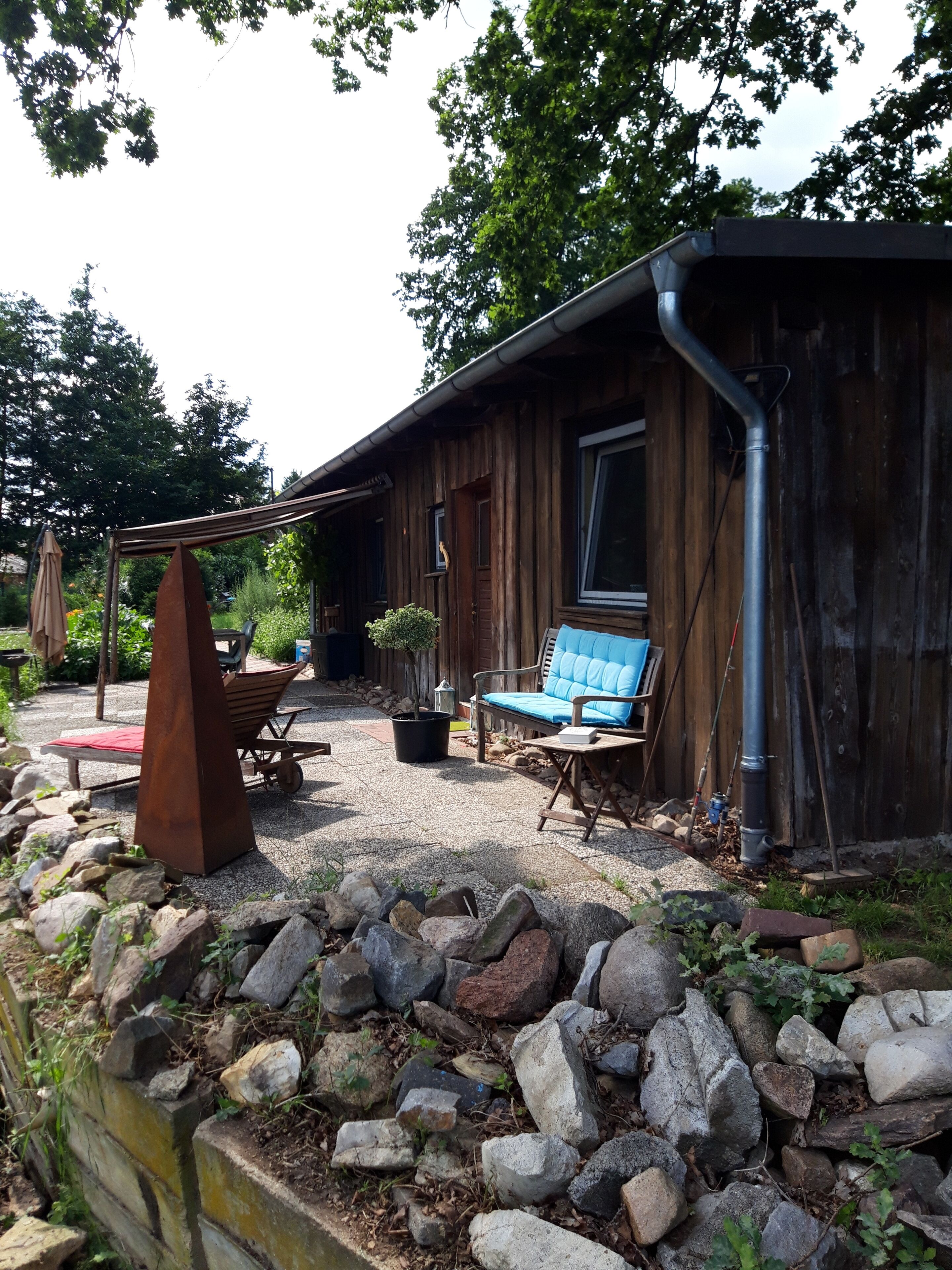 Une cabane en bois rustique dotée d’un patio comprenant un fauteuil bleu rembourré, une table en métal et une petite plante en pot.