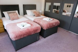 A hotel room with two beds, each with a pink blanket and white pillows. There are two bedside tables with lamps and a mirror on the wall. - Blue Apartment in Peterborough (Peterborough)
