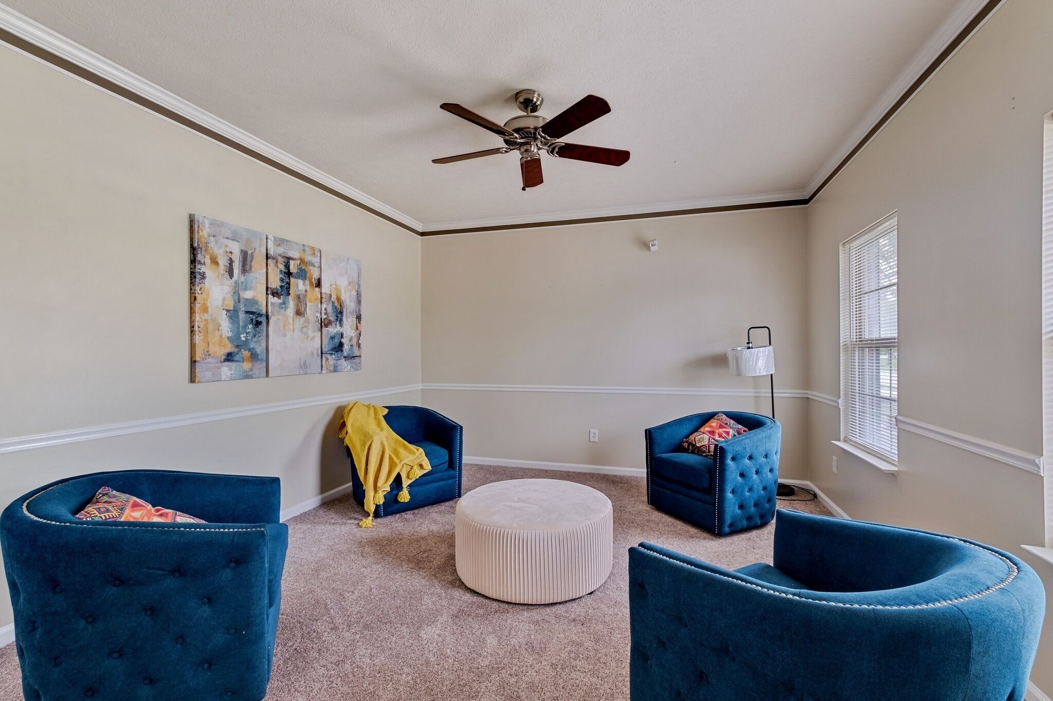 Une pièce dotée d’un ventilateur de plafond, de deux fauteuils bleus, d’un pouf rond et d’œuvres d’art abstraites accrochées au mur.