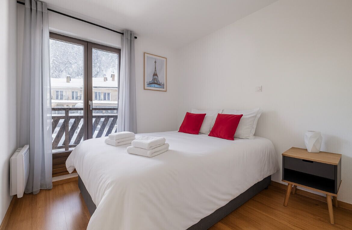 Hotelkamer met een groot bed, witte beddengoed, rode kussens, een nachtkastje en een raam met uitzicht op gebouwen.