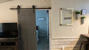 A room with a sliding door leading to a bedroom, a mounted TV, a wooden bench, and a wall-mounted mirror with a sign.