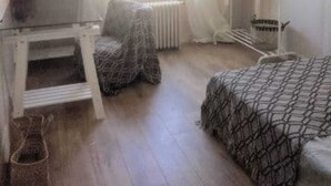 A small, sparsely furnished room with a wooden floor, a radiator, a window with curtains, a small table, a chair, a bed with a patterned blanket, and a clothing rack. - Location 31 Juillet au 15 Aout (Paris)