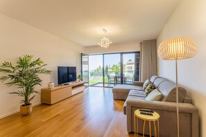 A modern living room with a sofa, TV, wooden floor, potted plant, and a balcony with a view.