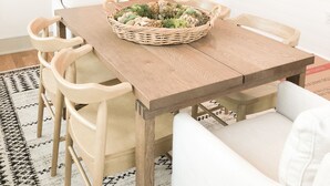 Un coin repas avec une table en bois, des chaises blanches, un luminaire en fibres tressées et un panier rempli de plantes vertes.