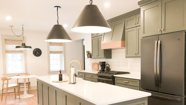 Une cuisine dotée d’un grand îlot central, de meubles de couleur vert foncé, d’appareils électroménagers en acier inoxydable, de luminaires suspendus et d’une fenêtre avec des stores.