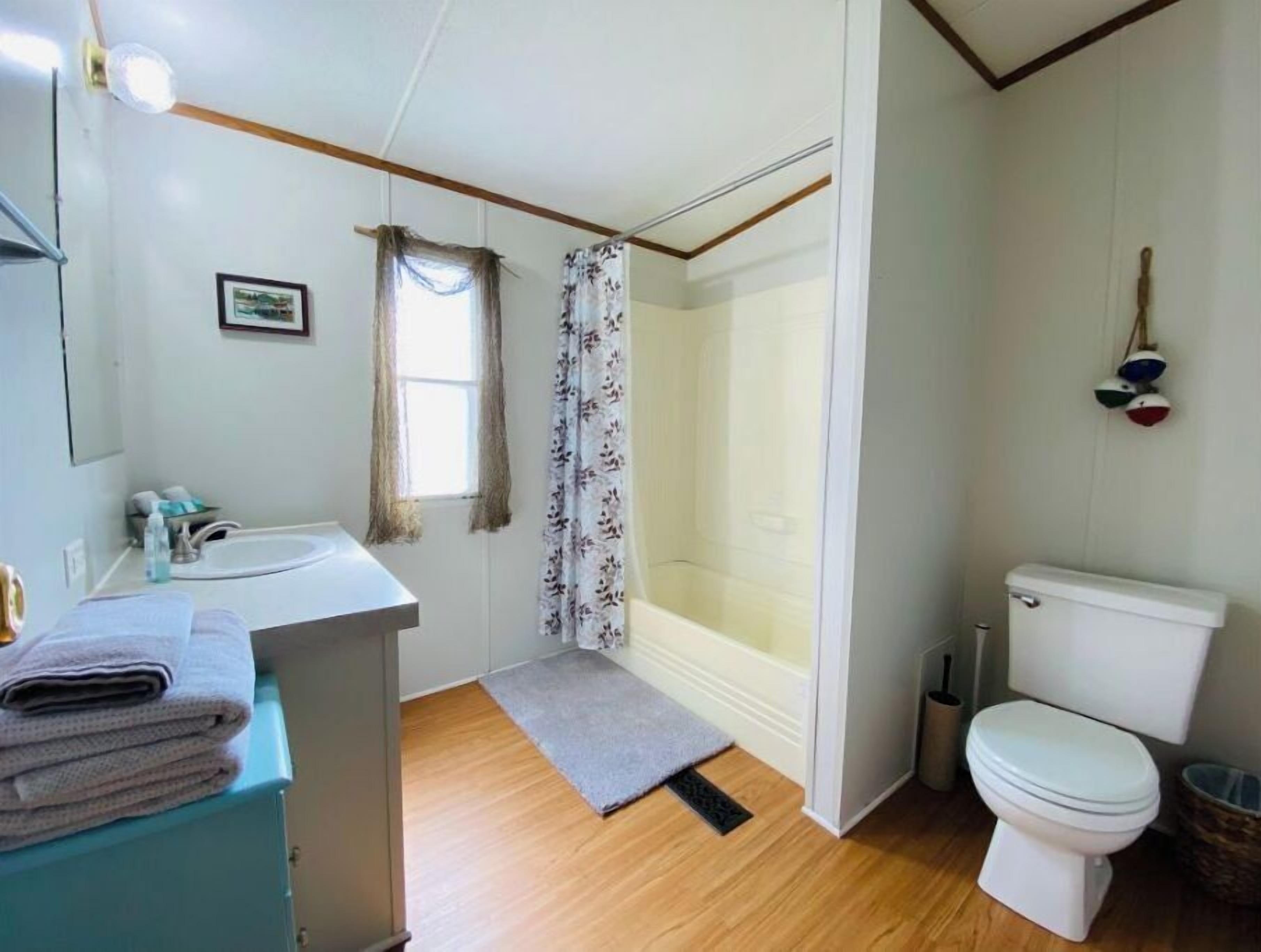 A bathroom with a white sink, toilet, and shower area. There is a window with curtains, a small rug, and a towel rack with towels.