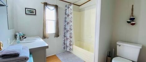 A bathroom with a white sink, toilet, and shower area. There is a window with curtains, a small rug, and a towel rack with towels.