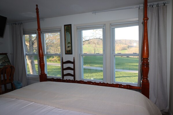 A bedroom with a four-poster bed, a wooden chair, and a view of a green landscape through large windows.