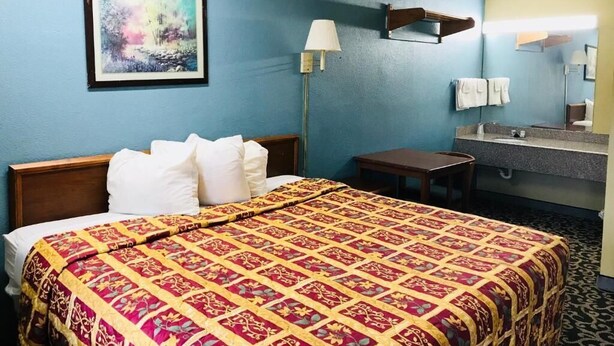 A hotel room with a large bed, a desk, a chair, a lamp, a painting, a mirror, and a bathroom with a sink and towel rack. - Roosevelt inn Motel (San Antonio)