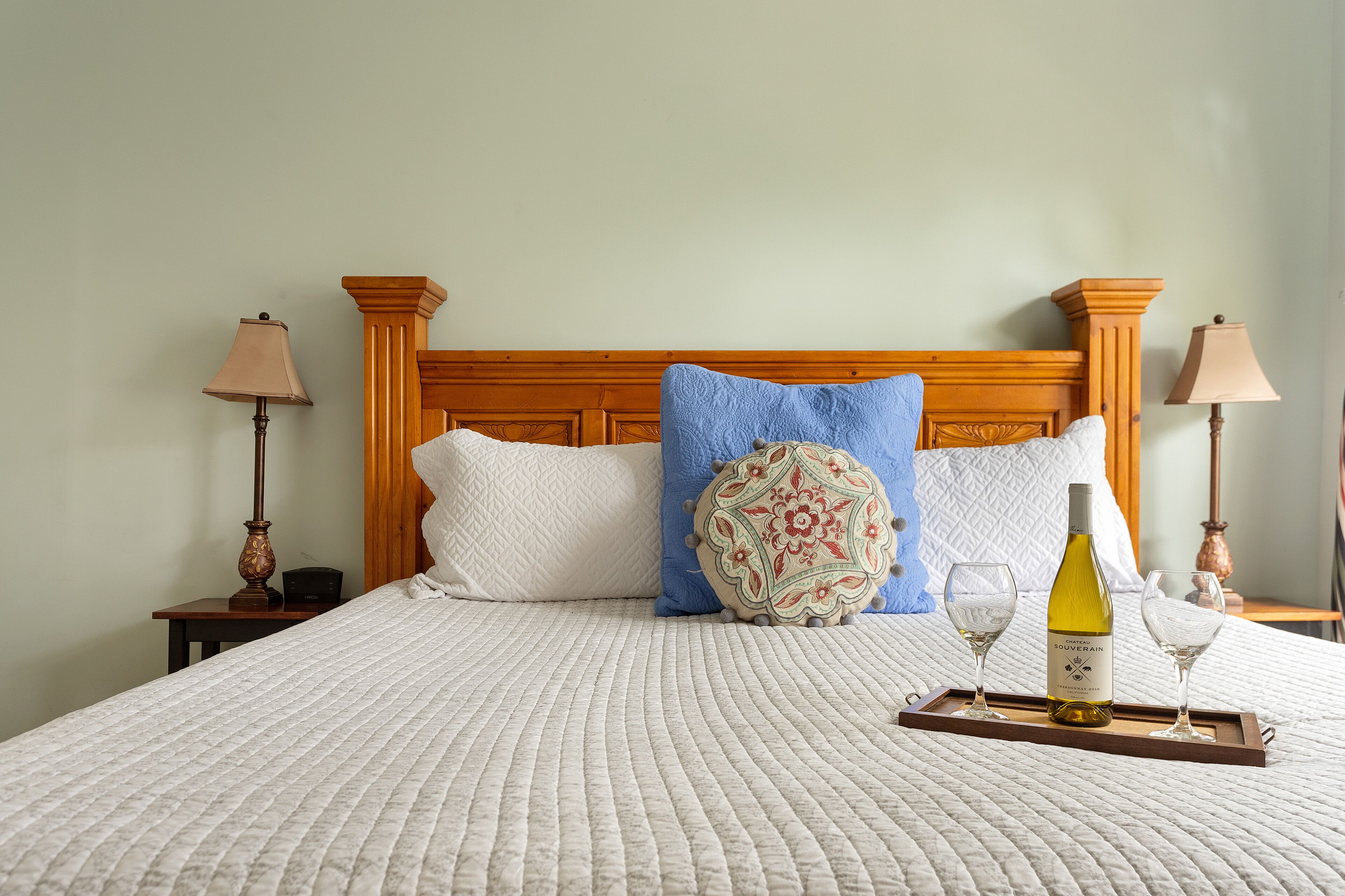 A neatly made bed with a quilted white cover, a decorative pillow, a tray with a wine bottle and glasses, and two bedside lamps.