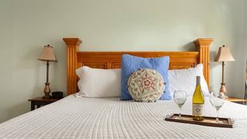 Cama bem arrumada com cobertura branca acolchoada, um travesseiro decorativo, uma bandeja com uma garrafa de vinho e copos, e duas luminárias de cabeceira.