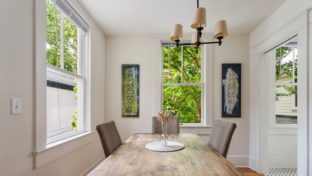A dining room with a wooden table, four chairs, a chandelier, and two windows with artwork.