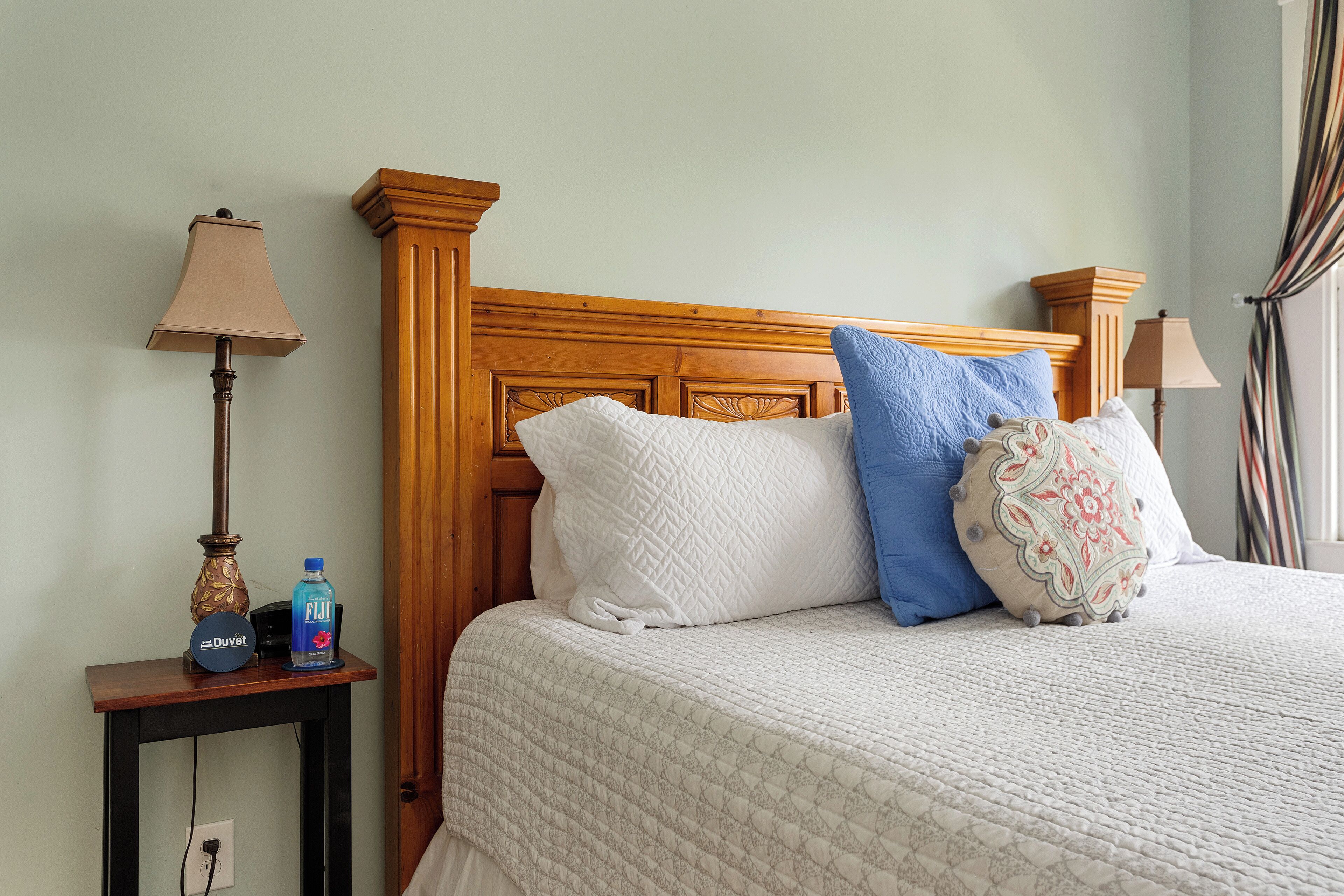 Una cama impecablemente arreglada con una colcha blanca acolchada, un cabecero de madera y almohadas decorativas. Una mesita de noche contiene una lámpara, una botella de Fiji y un cartel.