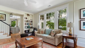 A spacious living room with a beige sofa, a wooden coffee table, and a side table with a lamp. There is a bookshelf filled with books and decorative items. - Downtown Luxury | The Rainbow Cottage (Charleston)