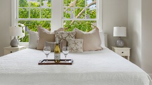 A neatly made bed with a wine bottle and glass, two bedside tables with lamps, and a window with a view of trees. - Downtown Luxury | The Rainbow Cottage (Charleston)
