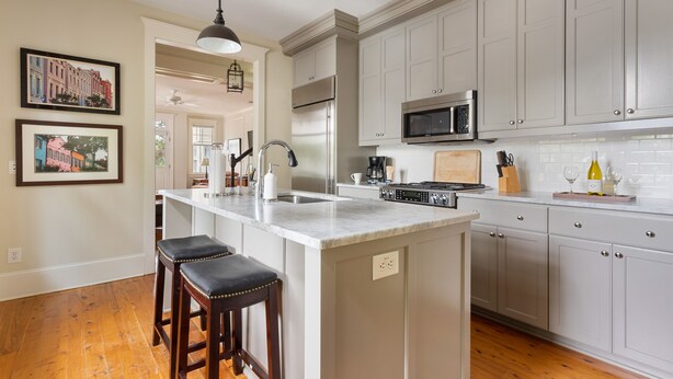 A kitchen with a large island, white cabinets, stainless steel appliances, and two bar stools. - Downtown Luxury | The Rainbow Cottage (Charleston)