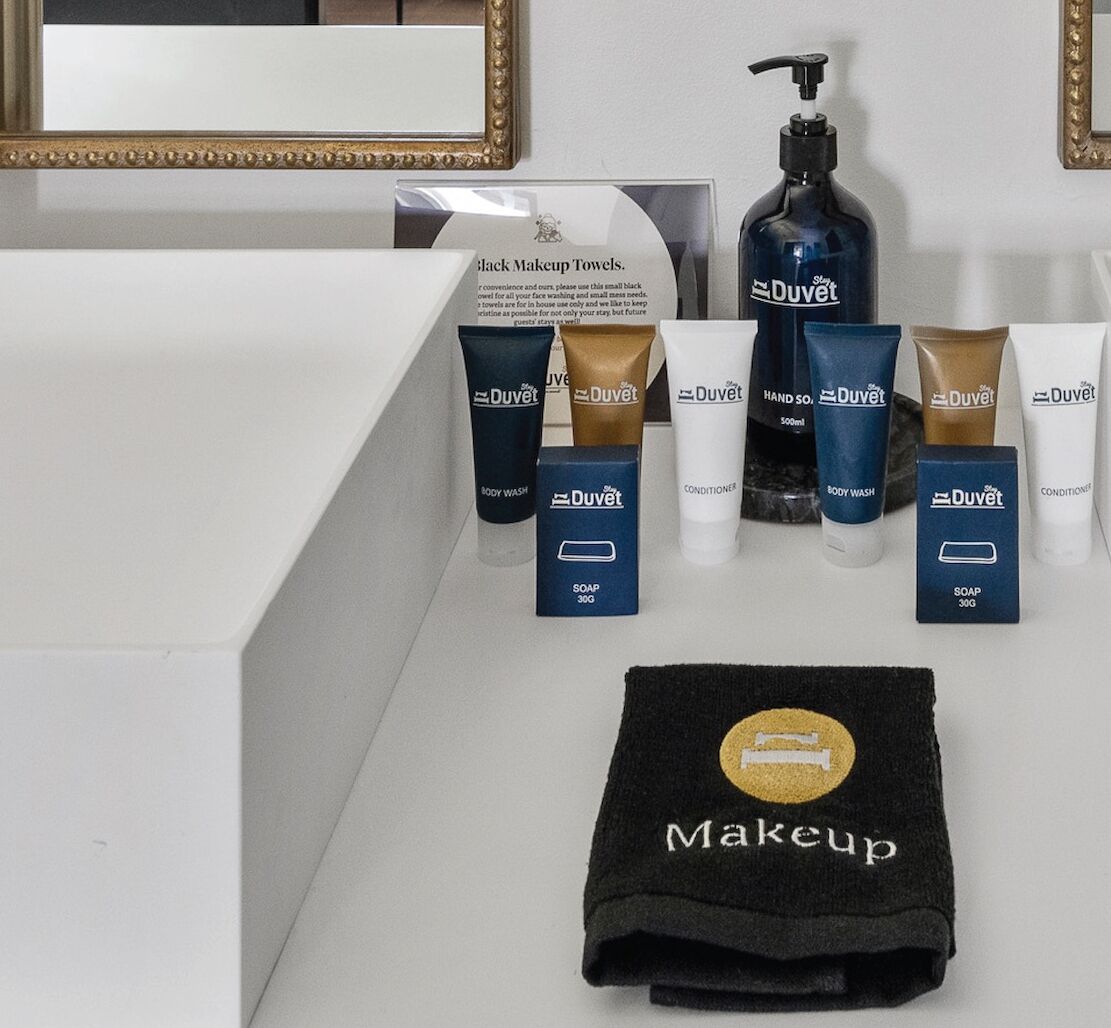 Ein Badezimmer-Waschtisch mit Spiegel, ein schwarzes Handtuch zum Abschminken sowie verschiedene Pflegeprodukte der Marke Duvet, darunter Duschgel, Conditioner und Seife.