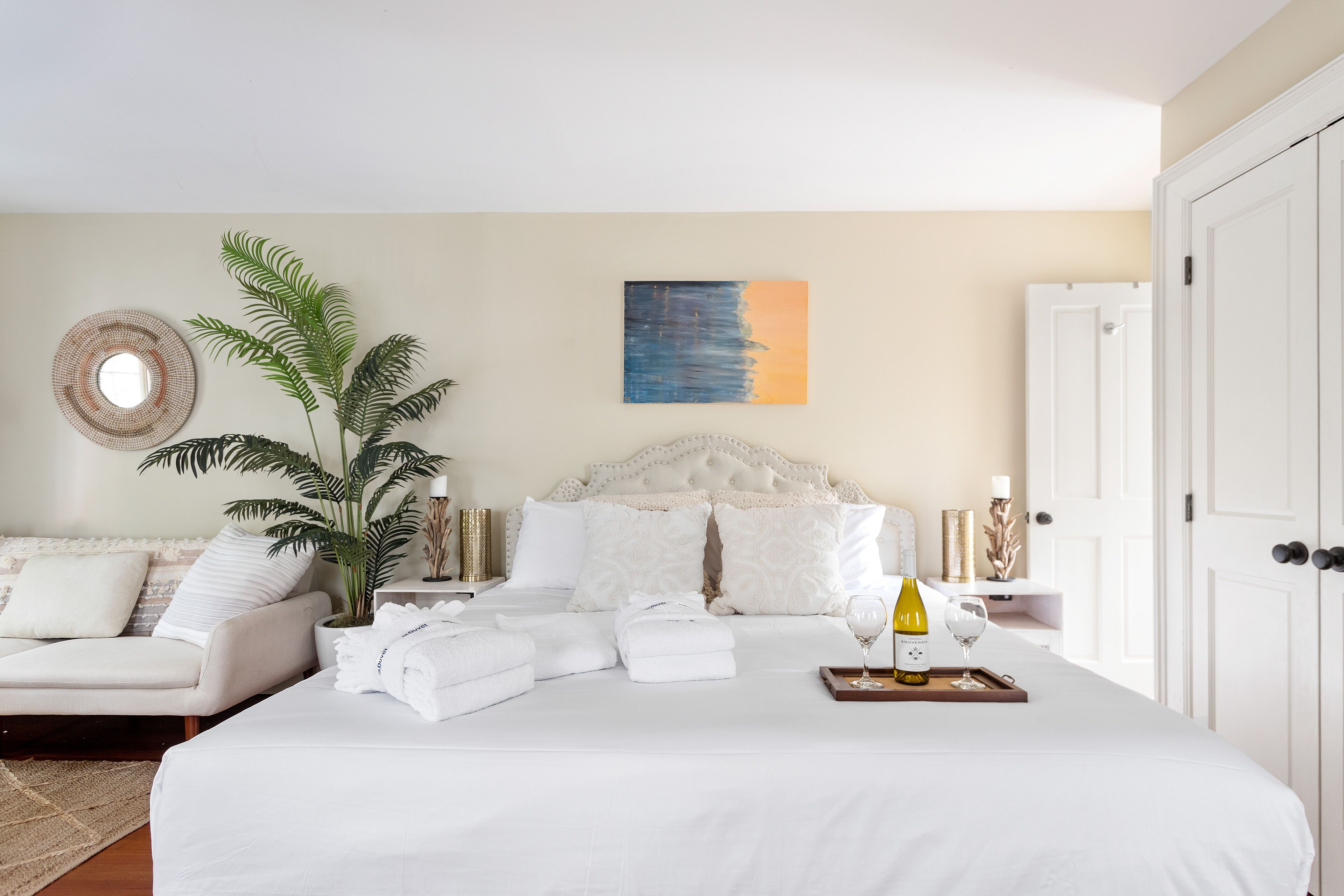 A neatly arranged bedroom with a large bed, a potted plant, a bedside table with a wine bottle, and a painting on the wall.