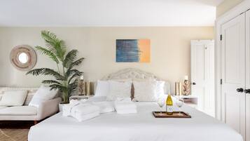 A neatly arranged bedroom with a large bed, a potted plant, a bedside table with a wine bottle, and a painting on the wall.