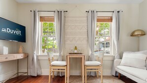 Una habitación con piso de madera, un sofá blanco, una pequeña mesa con dos sillas, un televisor montado en la pared y una ventana con cortinas.