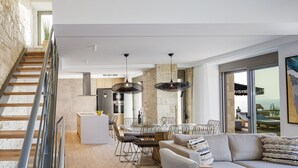 A spacious living area with a wooden staircase, a dining area with a round table and chairs, and a large window offering a view of the outdoors. - Perfect Hideaway for Families & Unmatched Sunsets (Rethymno)