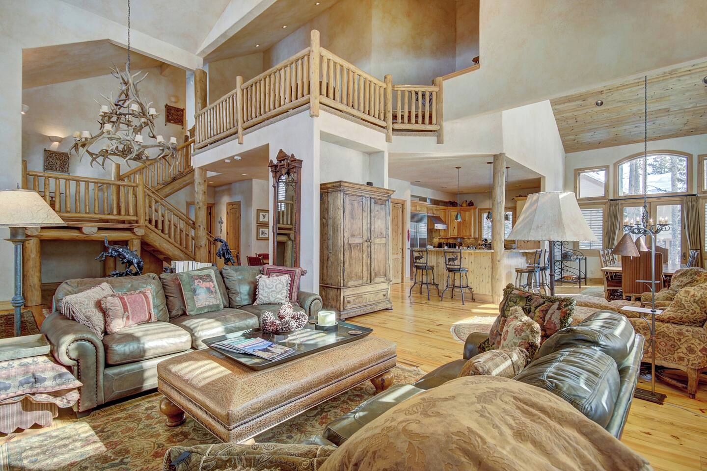 A spacious living area with a high ceiling, a grand staircase, and a kitchen in the background.