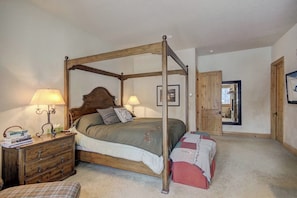 A four-poster bed with a wooden headboard, two bedside lamps, a nightstand with a lamp, a small bedside table, and a door leading to another room. - Windwood Breckenridge Luxury Mountain Home (Breckenridge)