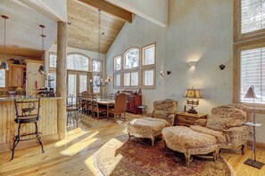 A spacious room with a high ceiling, wooden beams, a dining area with a large table and chairs, a cozy seating area with patterned ottomans, and a kitchen with wooden cabinets. - Windwood Breckenridge Luxury Mountain Home (Breckenridge)