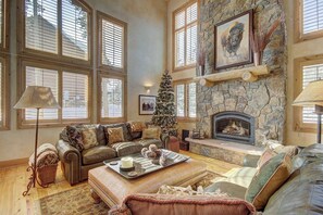 A cozy living room with a stone fireplace, a Christmas tree, and a large window with blinds. - Windwood Breckenridge Luxury Mountain Home (Breckenridge)