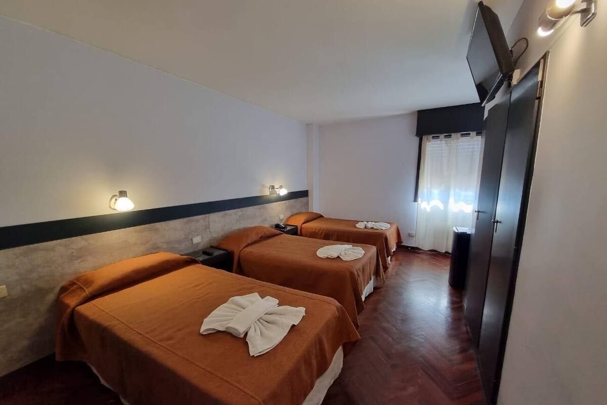 A hotel room with four beds, each with a brown bedspread and a towel. There is a television mounted on the wall and a trash can visible.