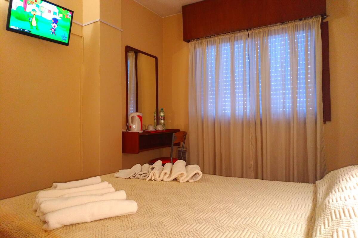 A hotel room with a bed, a television, a mirror, a window with curtains, and a small table with toiletries.
