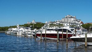 En småbåtshamn med flera yachter förtöjda vid träbryggor, en stor byggnad med en kupol och flera våningar i bakgrunden, och en klarblå himmel.