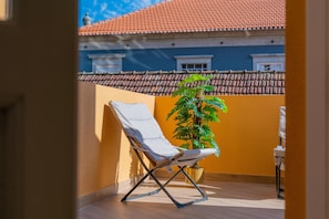 Ein Balkon mit weißem Stuhl, einem Blumentopf und Blick auf ein Gebäude mit rotem Ziegeldach.