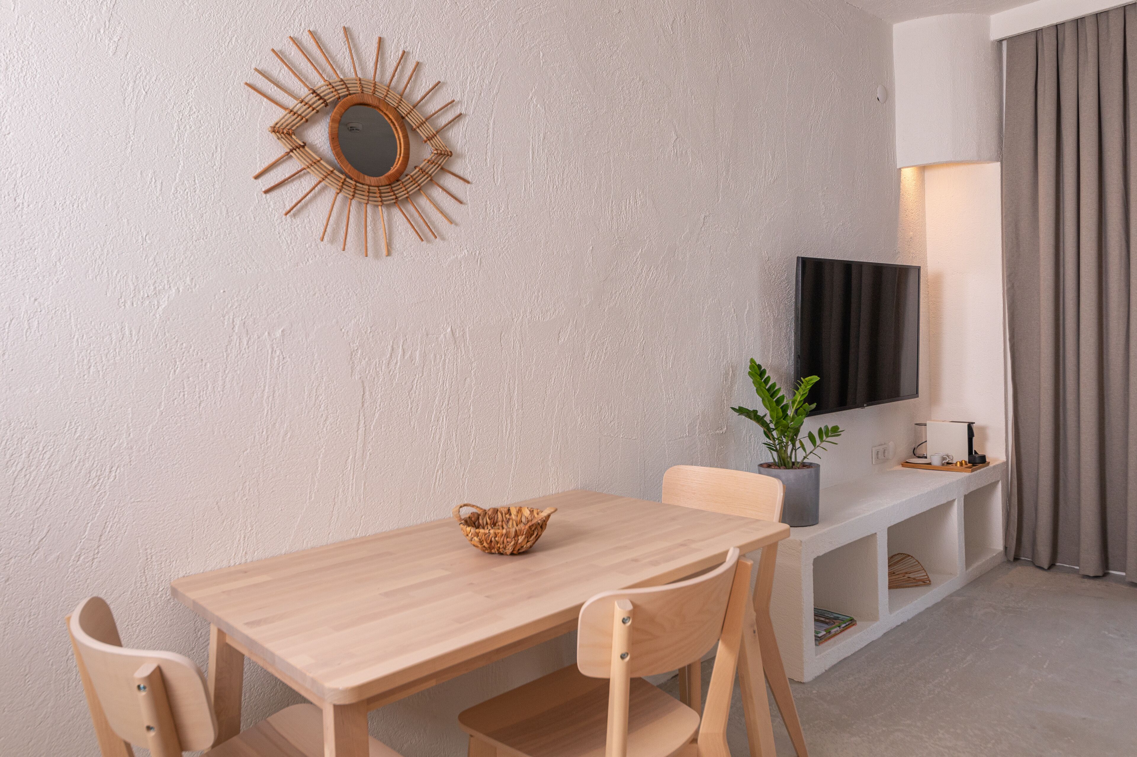 Un salon moderne avec une table en bois, deux chaises, un panier en osier, une plante en pot, un téléviseur à écran plat, une lampe et une grande fenêtre avec des rideaux.