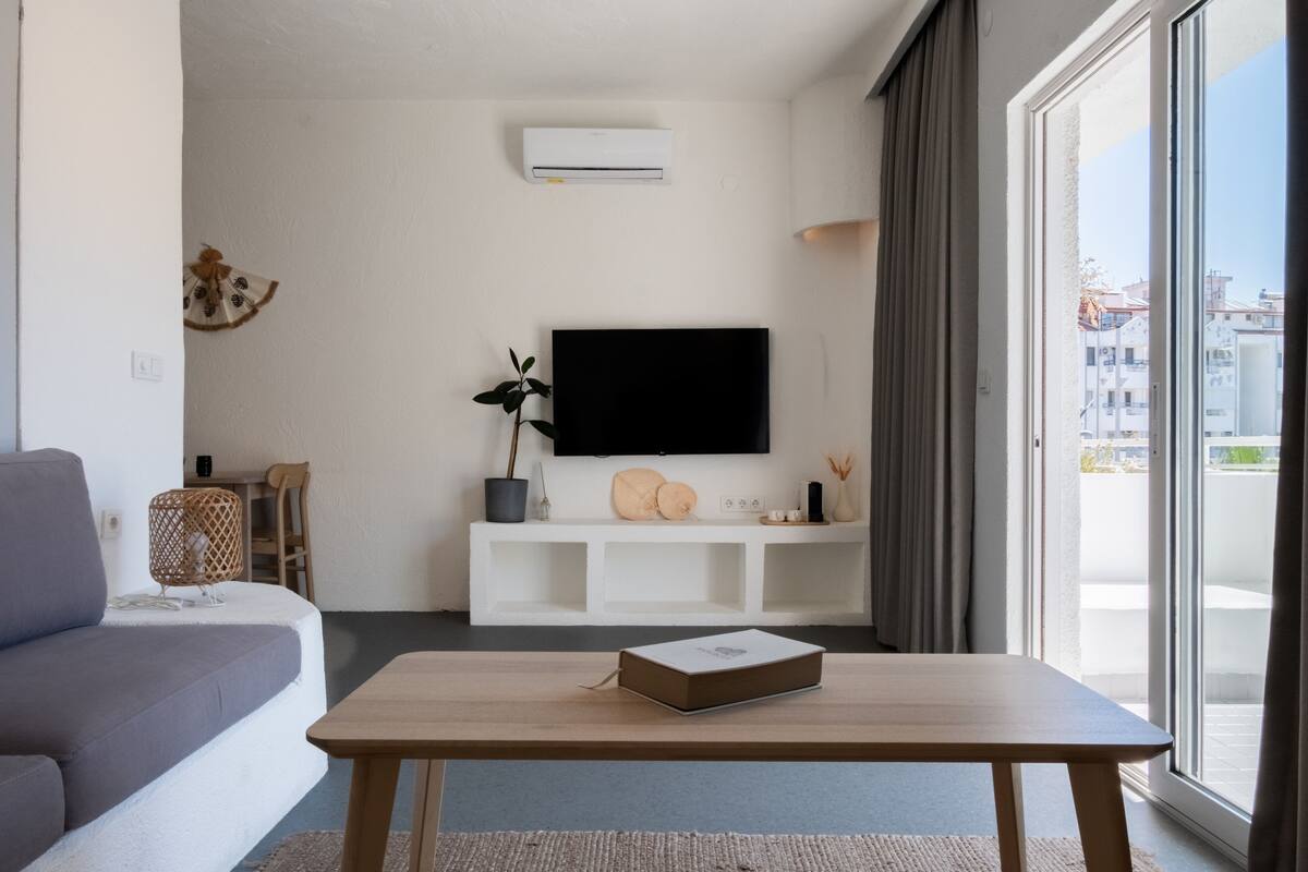 Un salon moderne avec un canapé gris, une table basse en bois, un téléviseur à écran plat posé sur une console blanche, une plante en pot et une vue sur les immeubles à travers de grandes fenêtres.