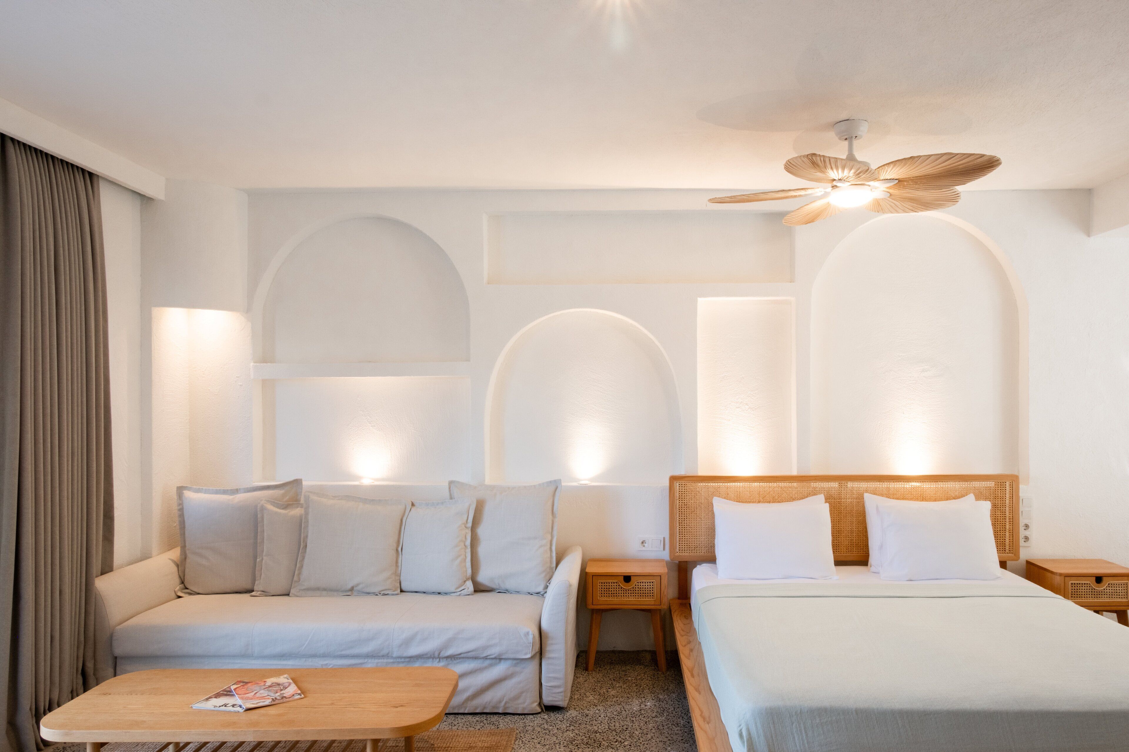 A hotel room with a bed, a sofa, a wooden nightstand, a table, and a ceiling fan.