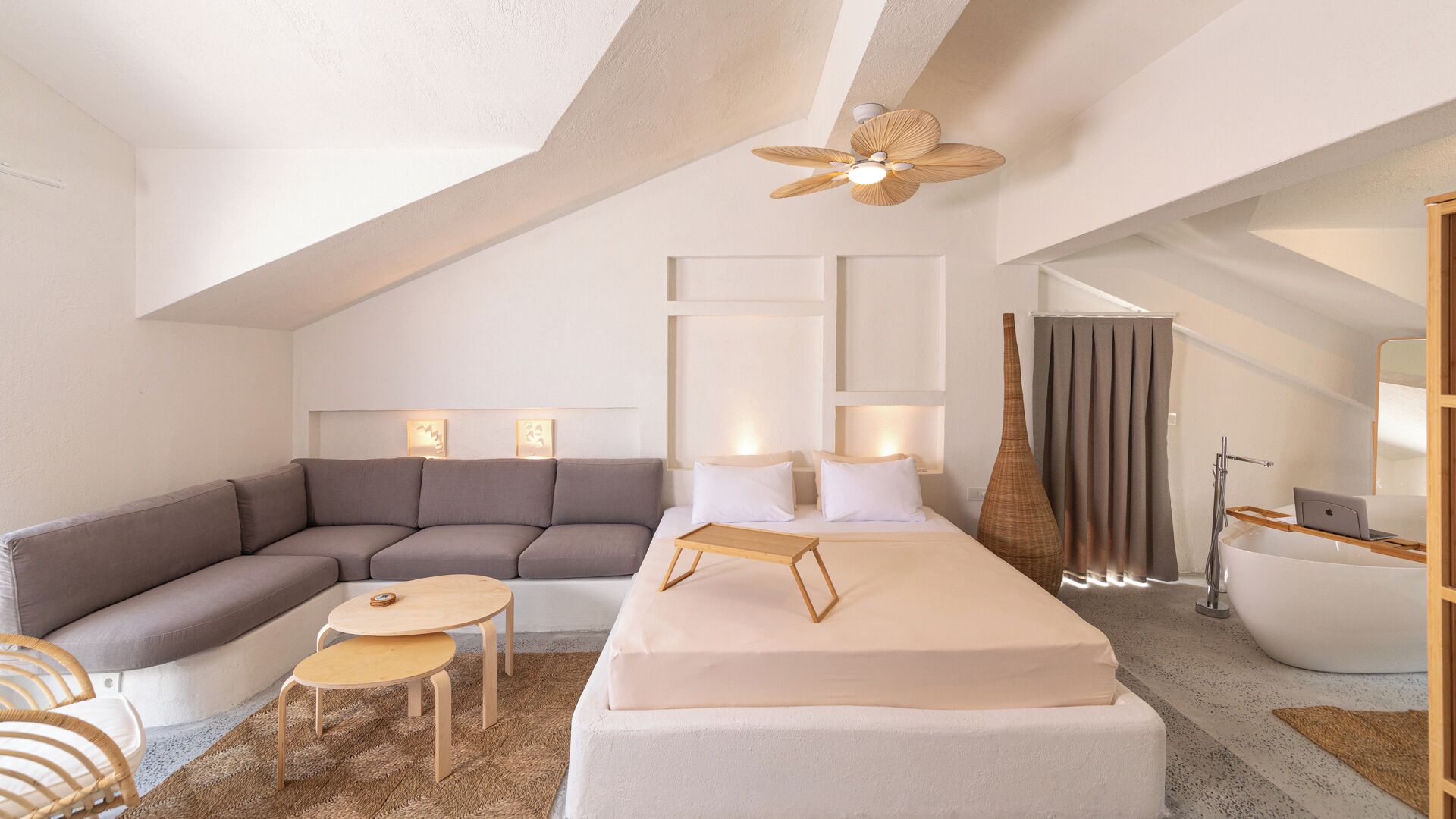 Un dormitorio tipo loft moderno y bien iluminado, con un sofá seccional gris, una cama con cabecera de madera, una pequeña mesa de madera y un ventilador de techo.