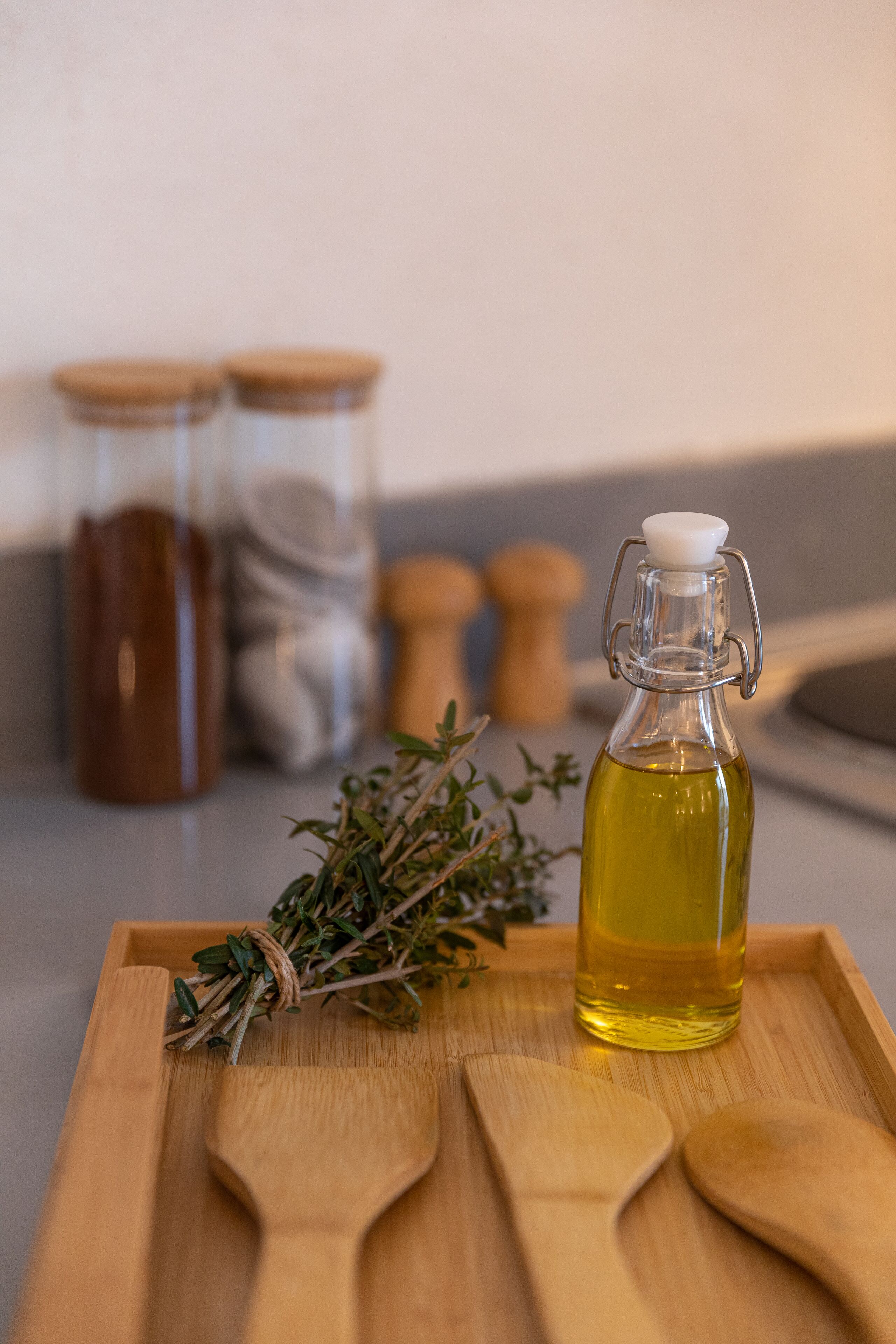 A wooden tray with a glass bottle of oil, two wooden spoons, and a bundle of herbs.