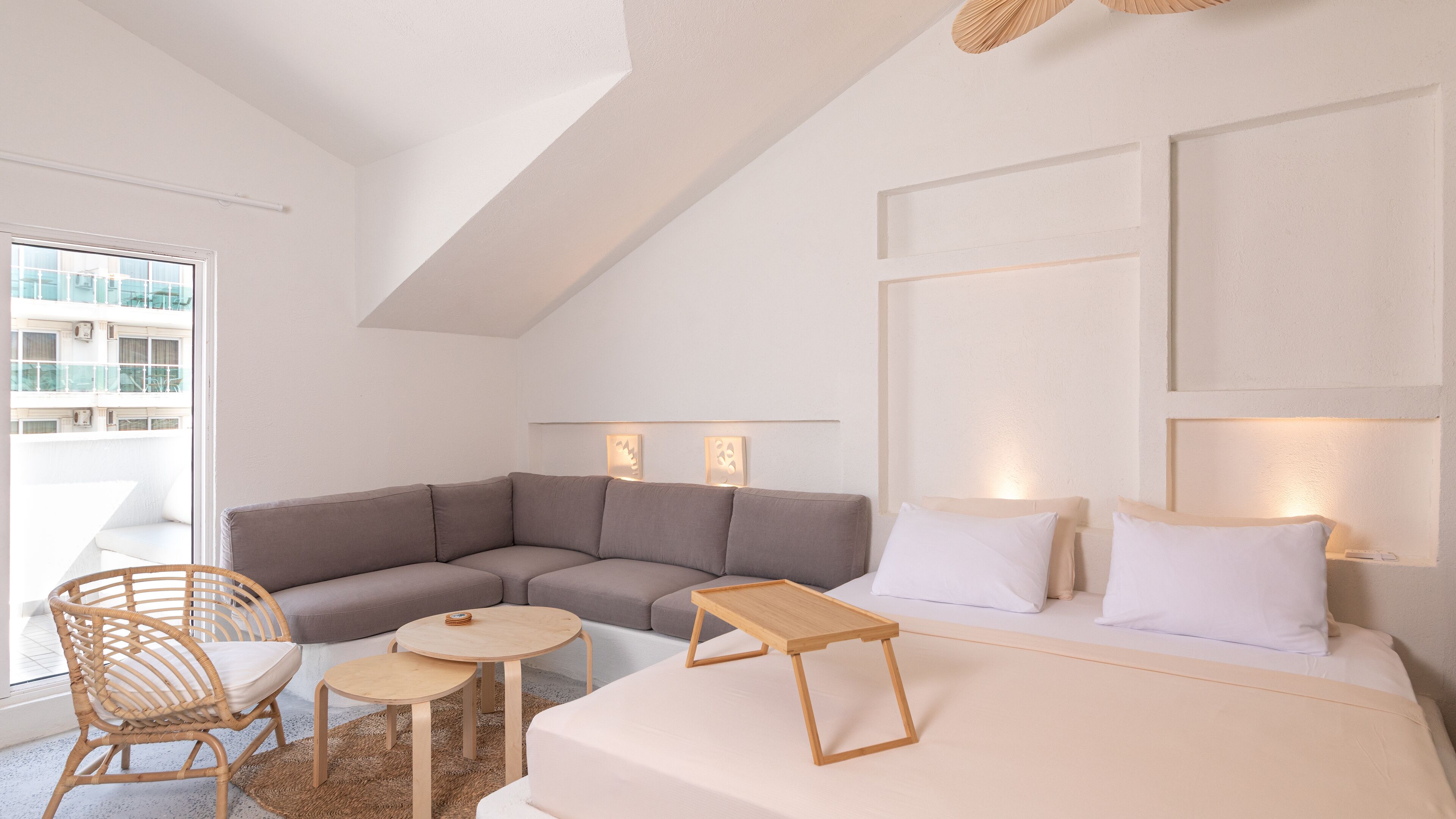 Una habitación de hotel moderna con una cama grande, un sofá seccional gris, una mesita redonda pequeña y una silla de mimbre.