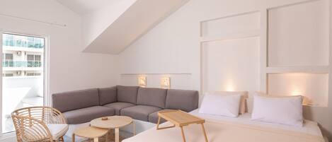 Una habitación de hotel moderna con una cama grande, un sofá seccional gris, una mesita redonda pequeña y una silla de mimbre.