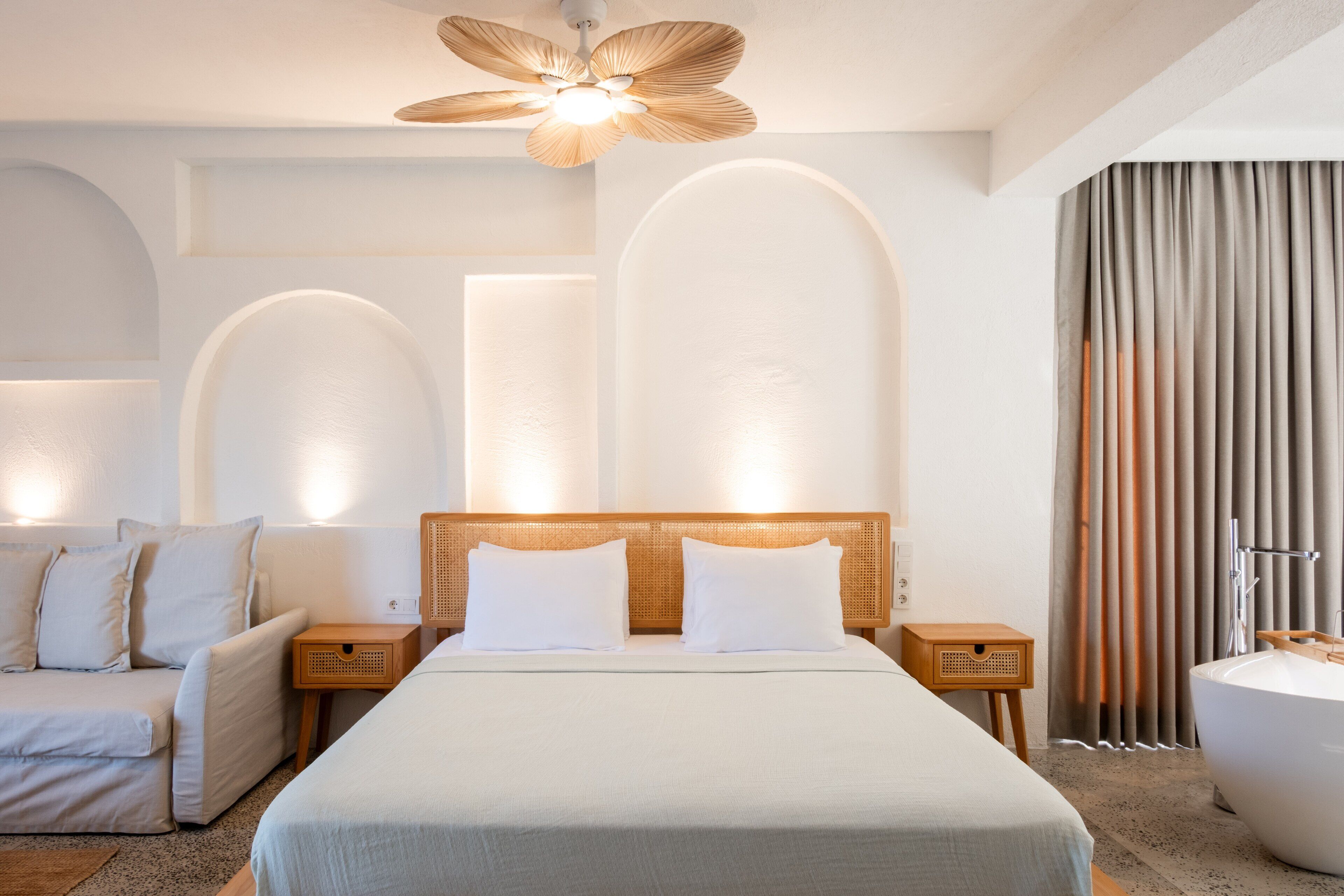 Quarto de hotel moderno com uma cama grande, um sofá, uma mesinha de cabeceira de madeira, um ventilador de teto e um banheiro com banheira.